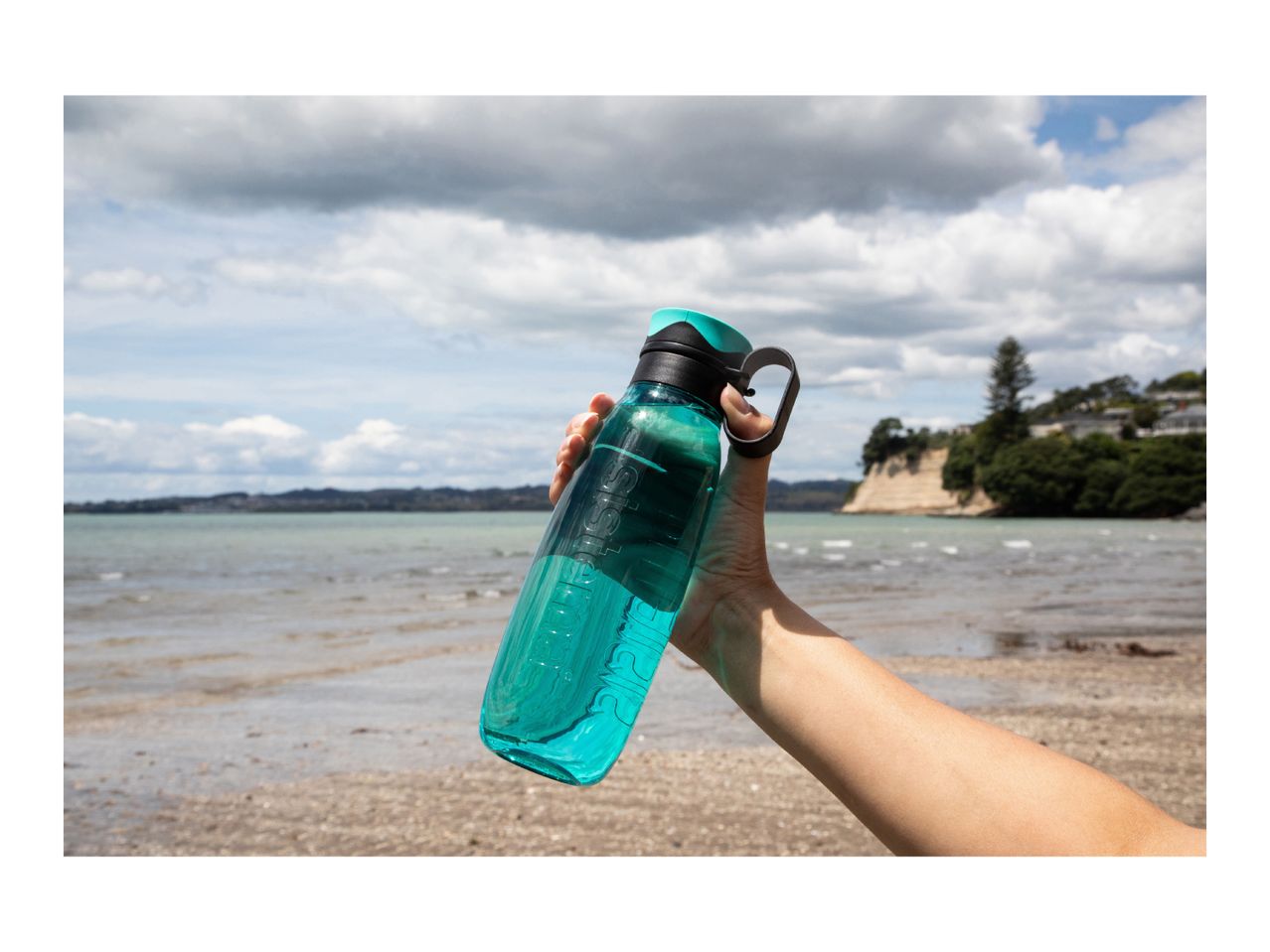 A hand holding a teal reusable water bottle at a beach.
