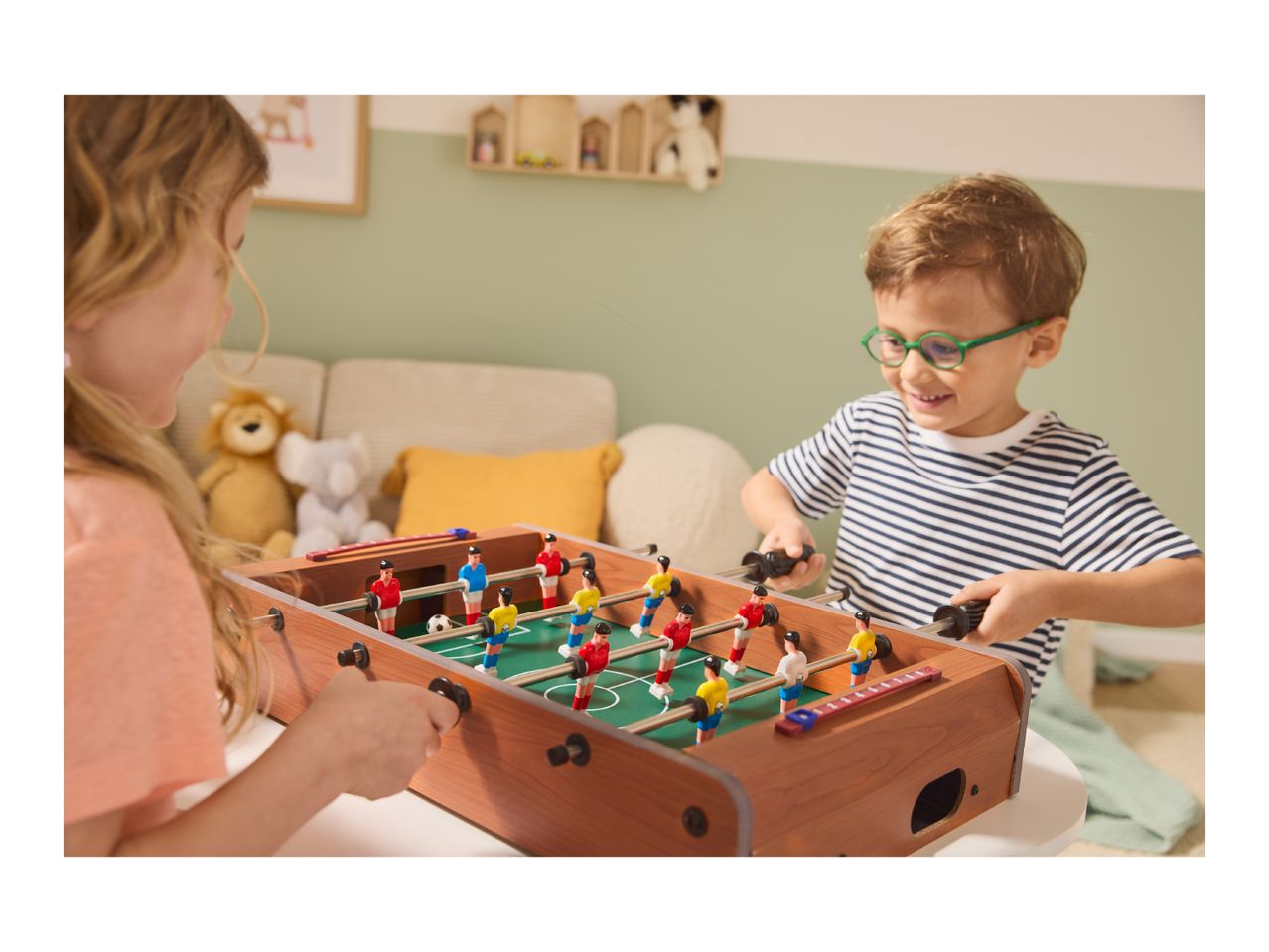 Two children playing foosball, a girl on the left and a boy in glasses on the right.