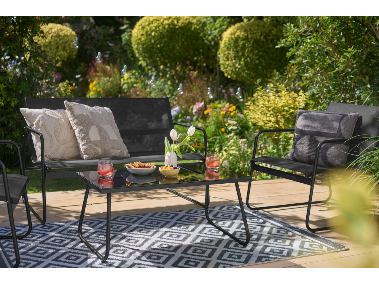 Crni baštenski nameštaj sa staklenim stolom, jastucima i grickalicama na terasi sa šarenim tepihom.