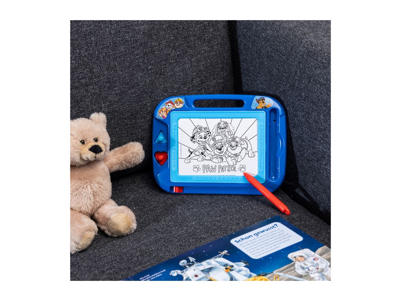 A blue Paw Patrol magnetic drawing board with a red pen, a teddy bear, and a book.
