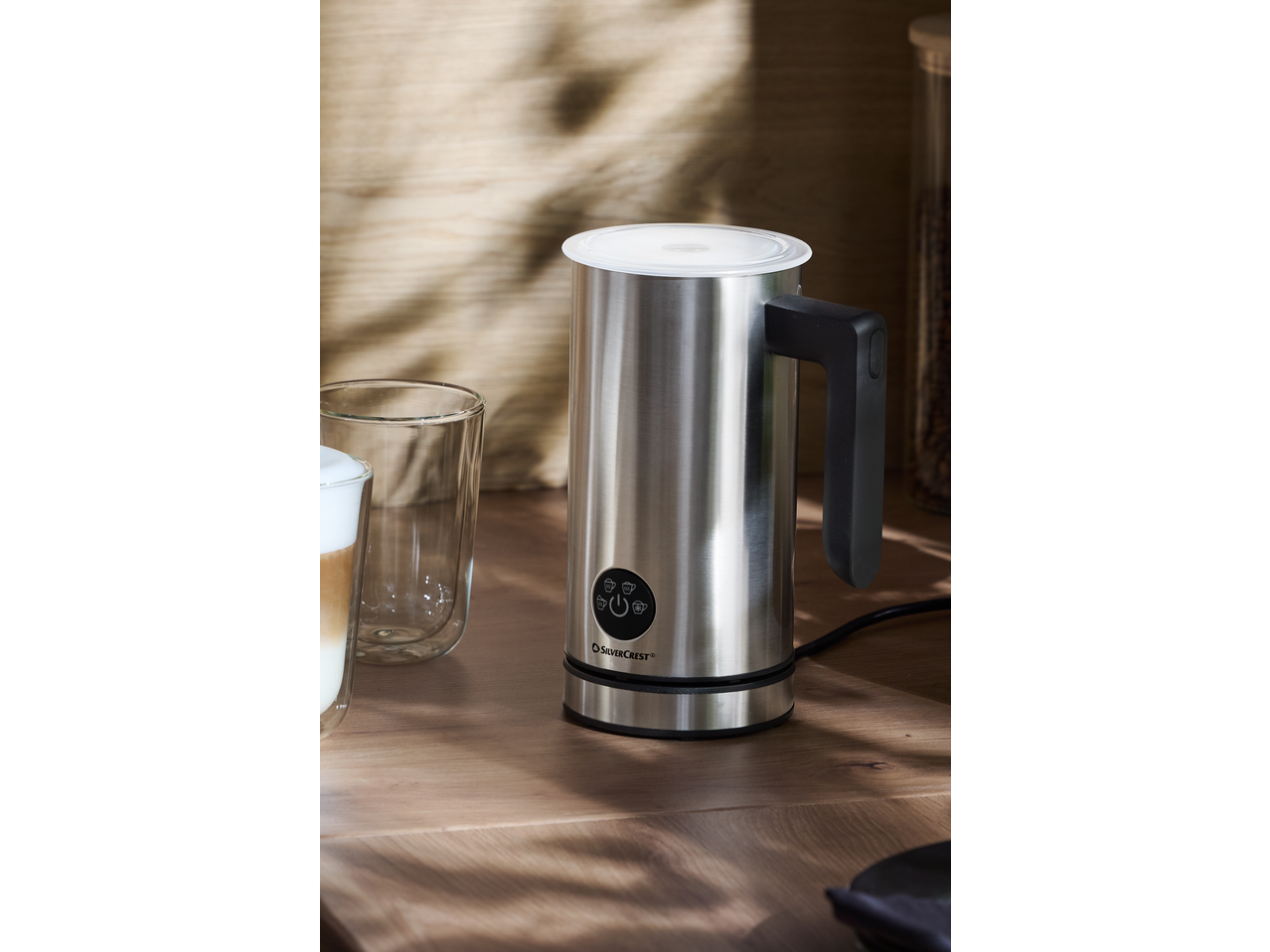 Stainless steel milk frother on a wooden counter next to a glass of latte.