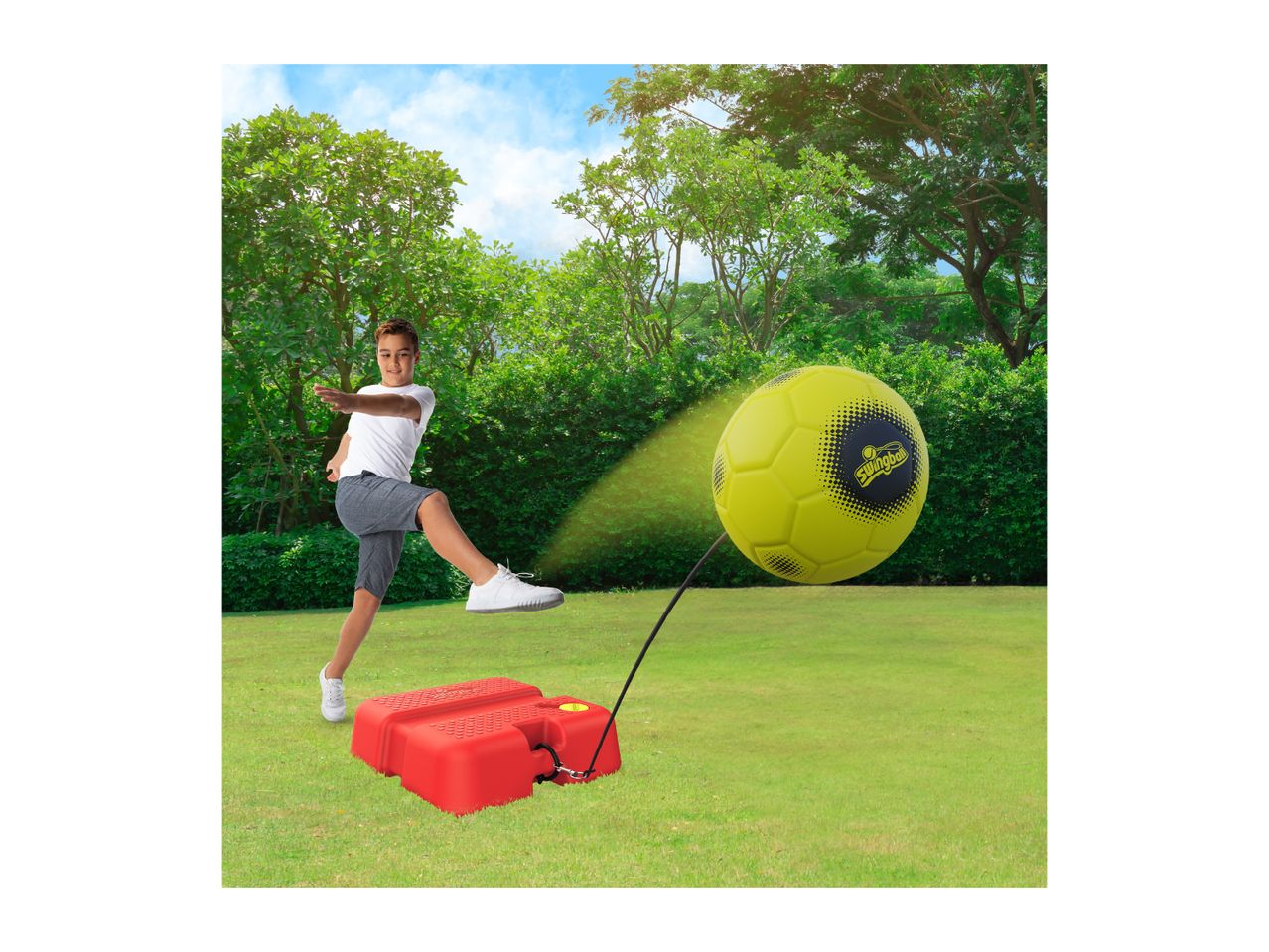 Boy kicking a yellow tethered football on a red base in a grassy garden.