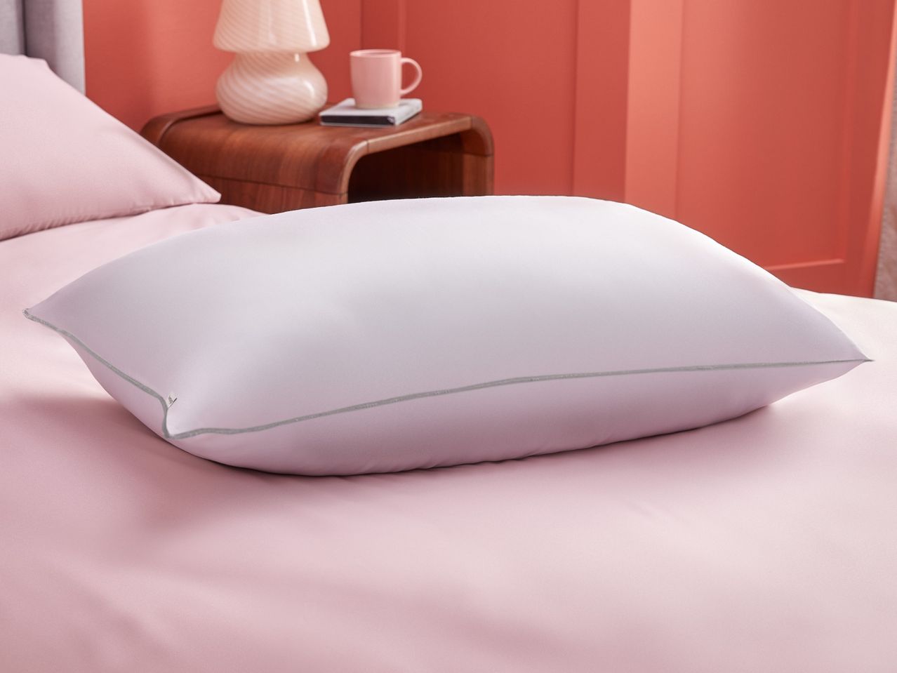 A light purple pillow sits on a pink bedsheet, next to a wooden bedside table.