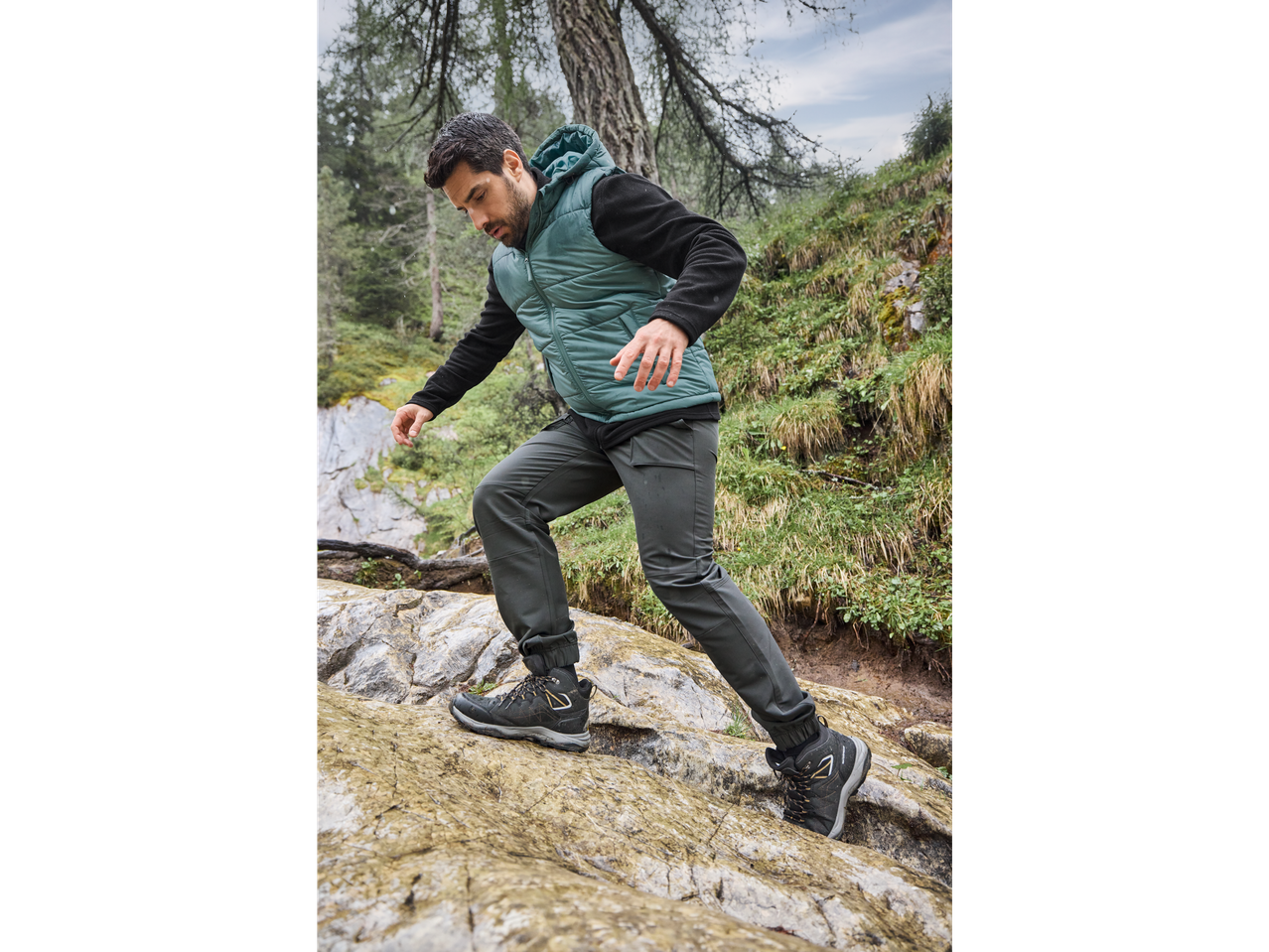 Man in a teal thermal sports gilet and dark pants hiking on a rocky trail.