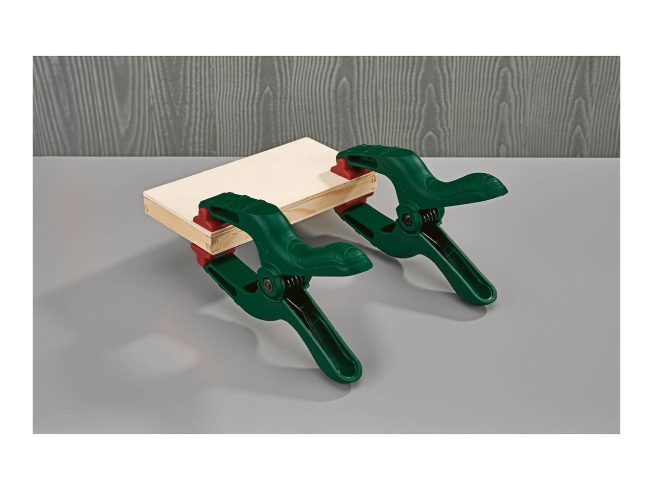Two green spring clamps holding a piece of light wood on a grey surface.