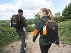 Two hikers with black and orange backpacks walk on a dirt path overlooking a lake.