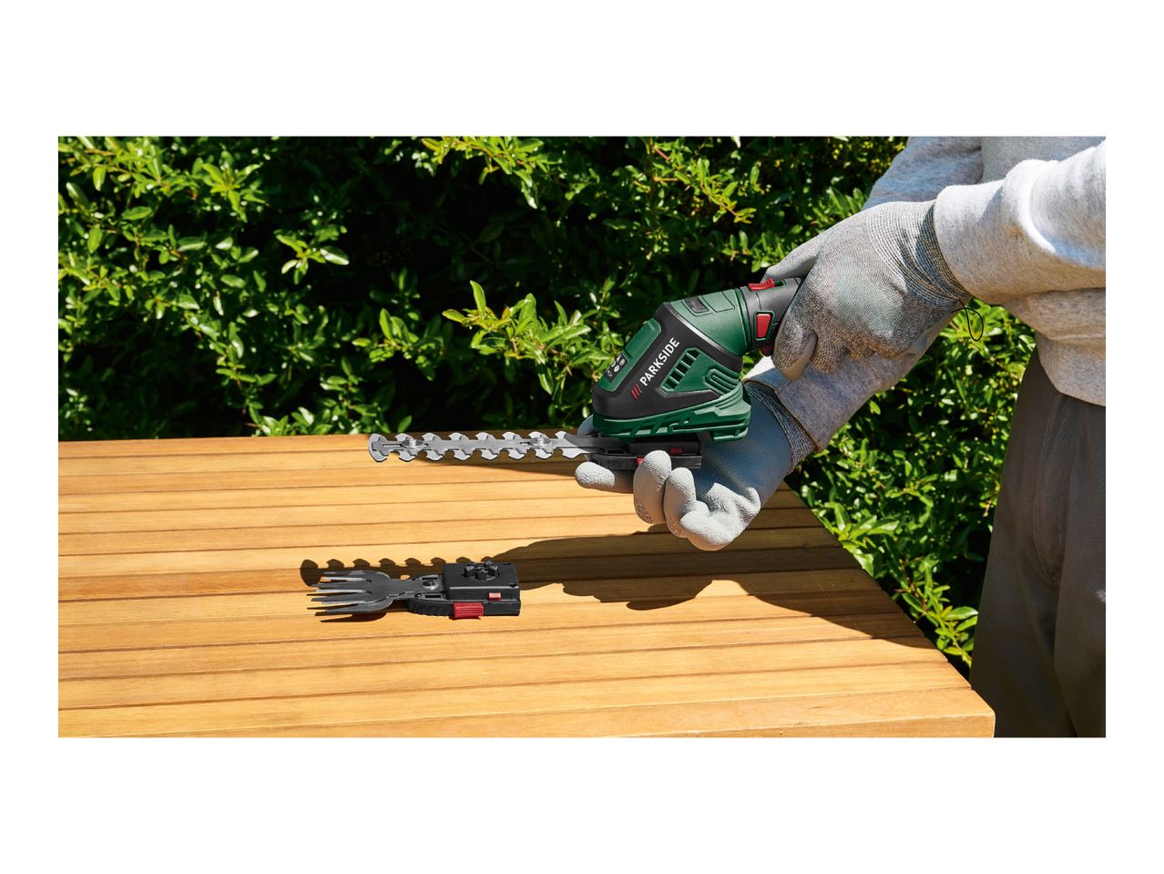 Person in gloves holding a hedge trimmer with another attachment on a wooden table.