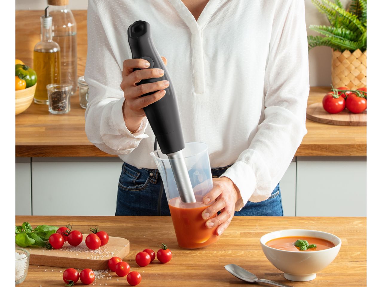 Woman blending tomato soup with an immersion blender in a kitchen setting.
