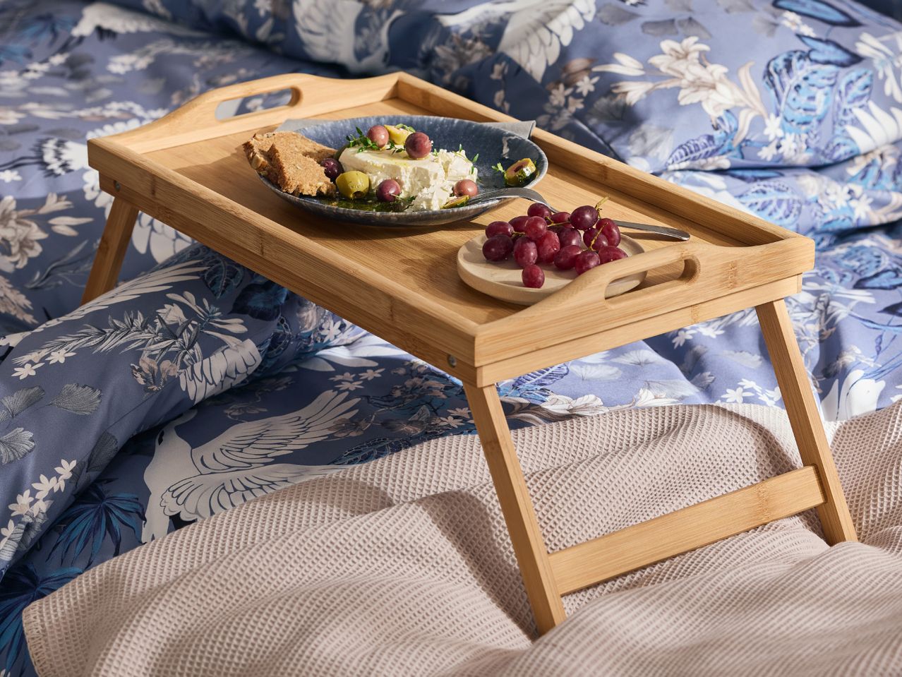 Breakfast in bed with a wooden tray holding feta cheese, olives, bread, and grapes.