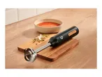 Black immersion blender on a wooden board with a bowl of tomato soup and herbs.