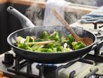 Steaming broccoli and other vegetables stir-fry in a dark-colored frying pan on a gas stove.