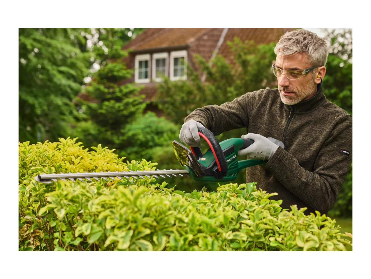 A man in safety glasses and earplugs trims a hedge with a hedge trimmer.