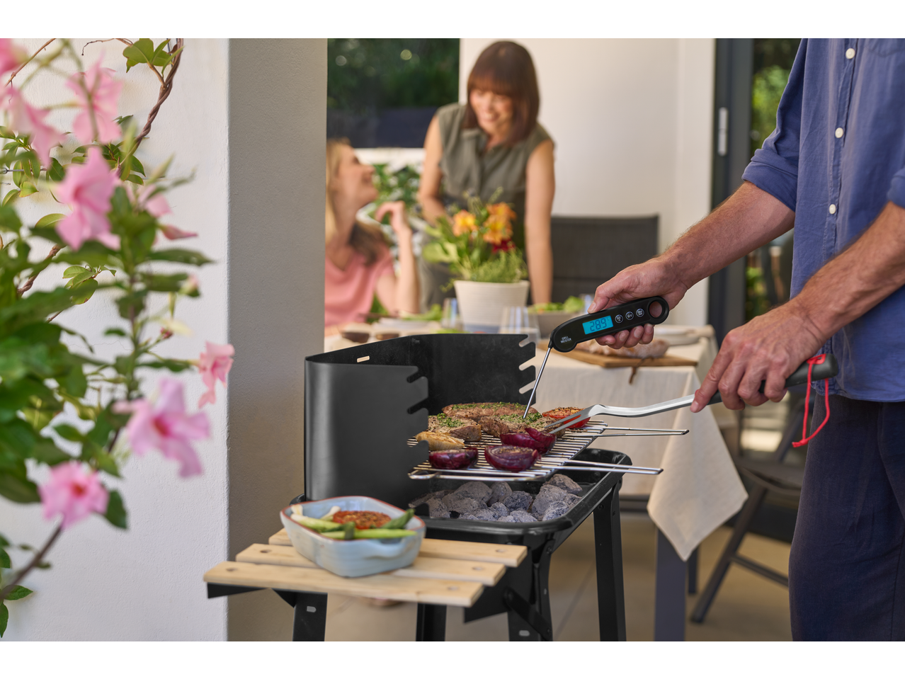 Bărbat măsoară temperatura cărnii pe grătar, femei la masă în fundal.