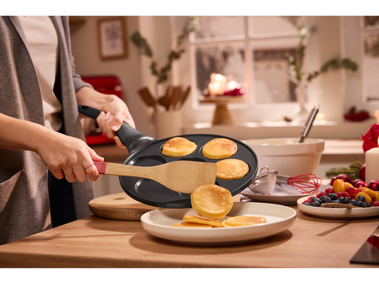 Žena priprema male palačinke sa Silvercrest® tiganjem, voće i sveće u pozadini.